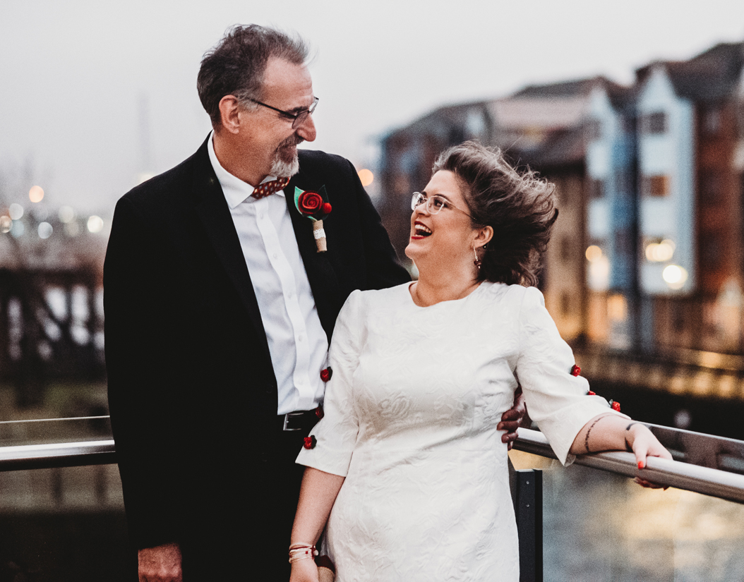 Norm Tovey-Walsh and Bethan Tovey-Walsh on their wedding day. He is wearing a suit, she's wearing a white dress. They are outside on a balcony with buildings and water behind them, and the wind is blowing their hair.
