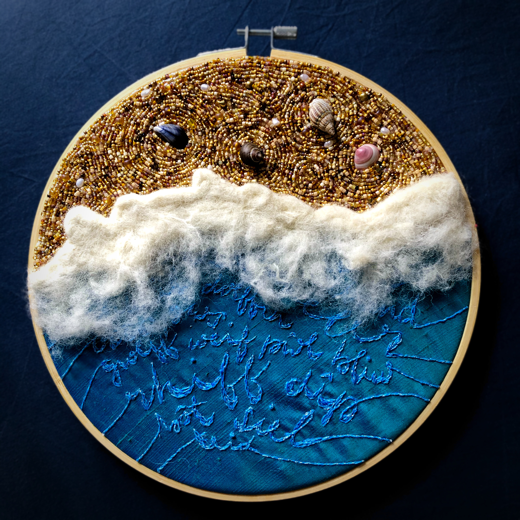 An embroidery hoop containing a representation of the sea washing over a sandy beach. The sand is composed of hundreds of glass and pearl beads. The sea is a piece of blue-green silk, upon which English loanwords into Welsh are embroidered.