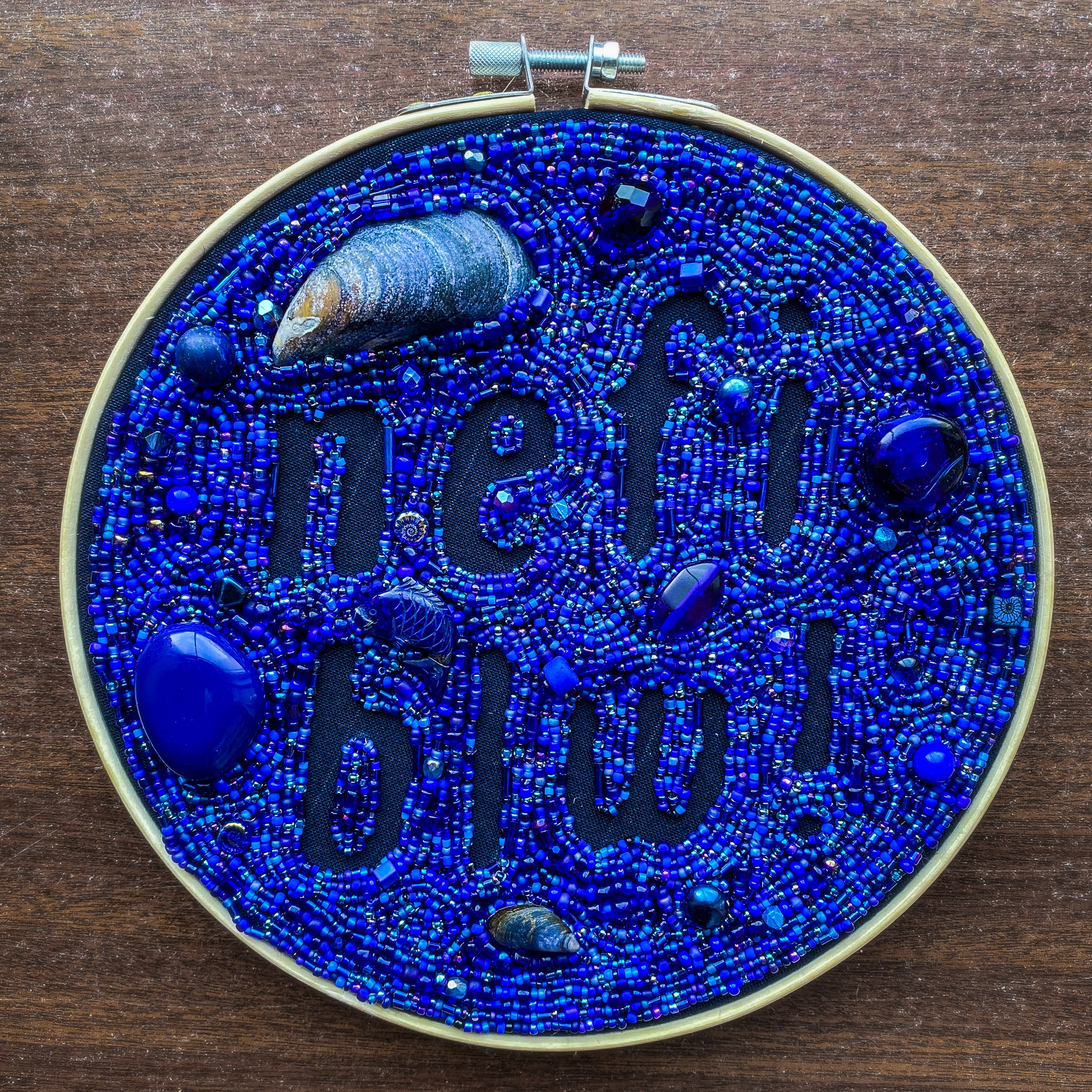 A round embroidery hoop containing a piece of navy blue fabric. The fabric is almost entirely covered with small beads and mussel shells, in various shades of dark blue. The spaces without beads form the Welsh words 'nefi blw'.
